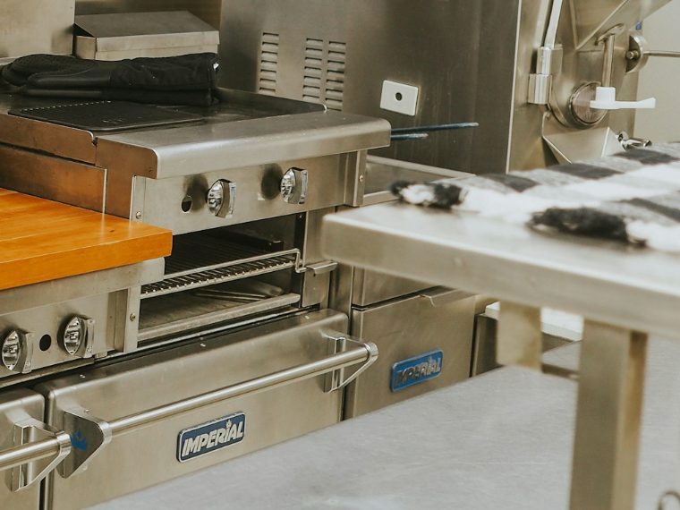 Commercial kitchen equipment — a gas range with an overhead salamander broiler beside a stainless steel prep table
